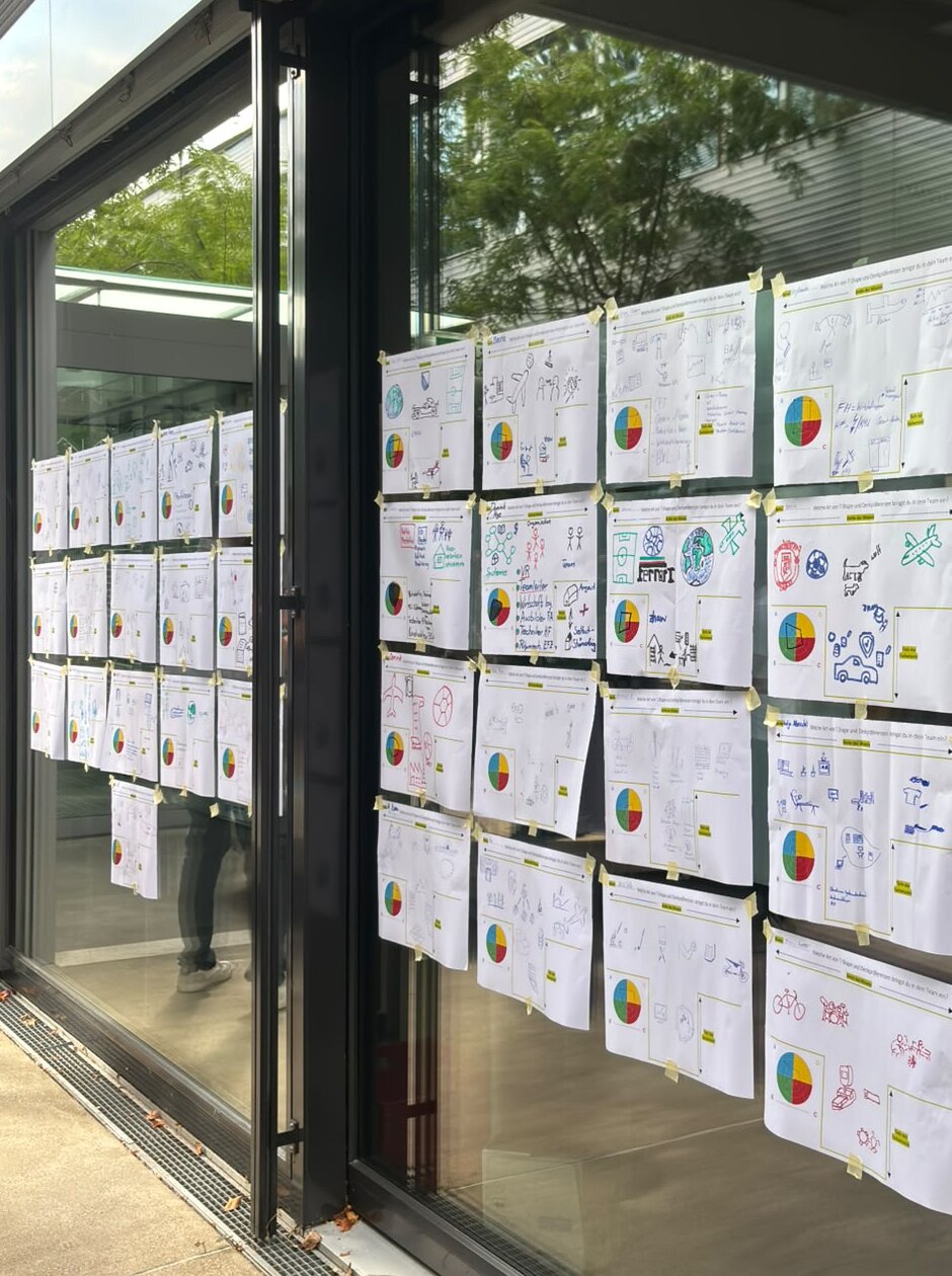 Dutzende handgezeichnete T-Shape-Plakate mit farbigen Kreisdiagrammen kleben mit Kreppband an einer Glasfassade.