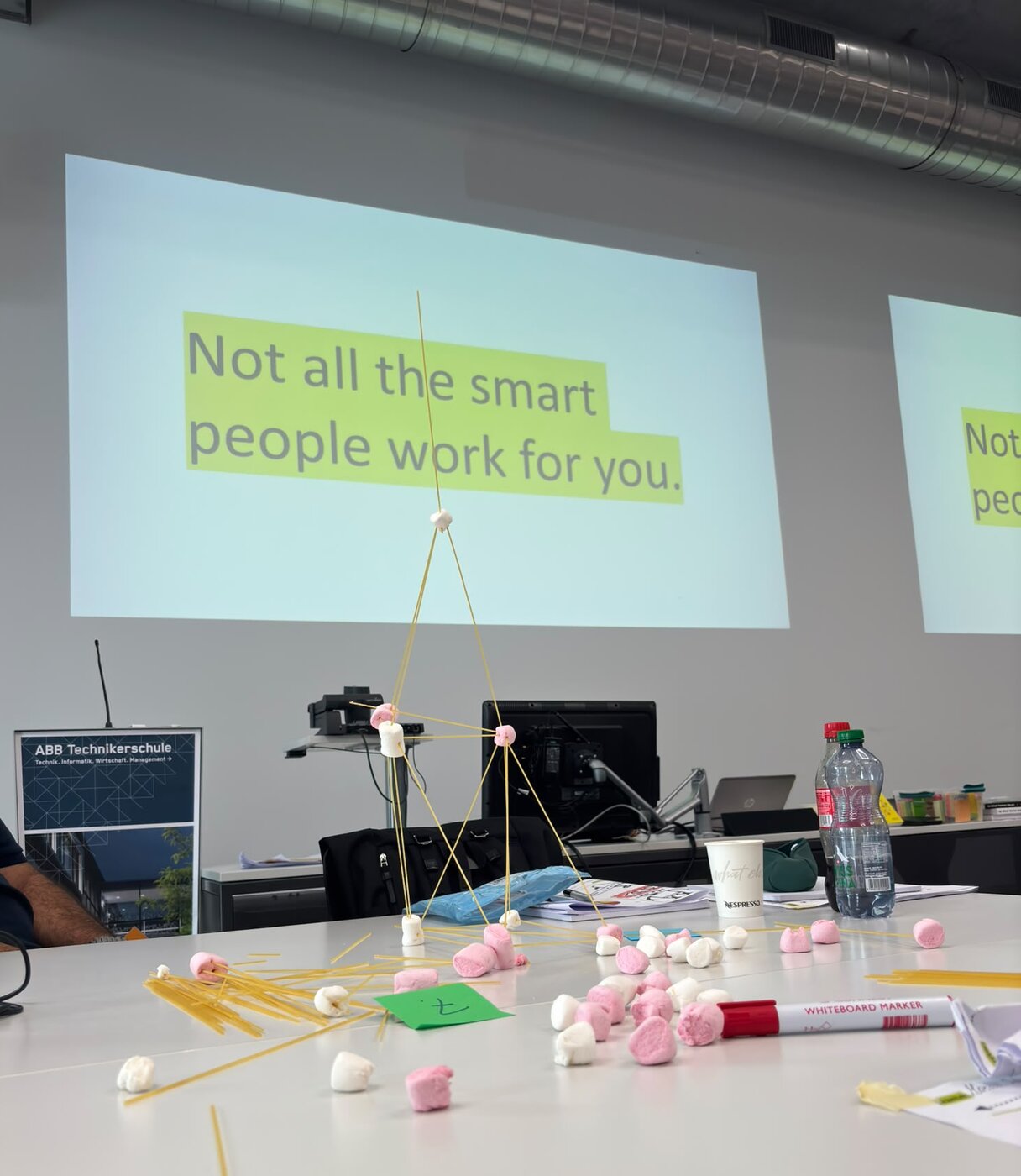 Ein Turm aus Spaghetti und Marshmallows steht auf einem Tisch, dahinter die Leinwand mit dem Satz Not all the smart people work for you.
