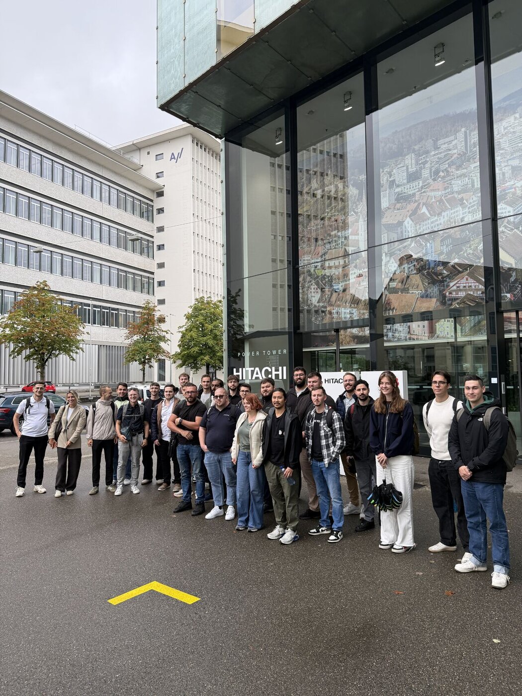 Die Gruppe der Summer-School-Teilnehmenden steht vor dem verglasten Eingang des Power Tower mit dem Schriftzug Hitachi.
