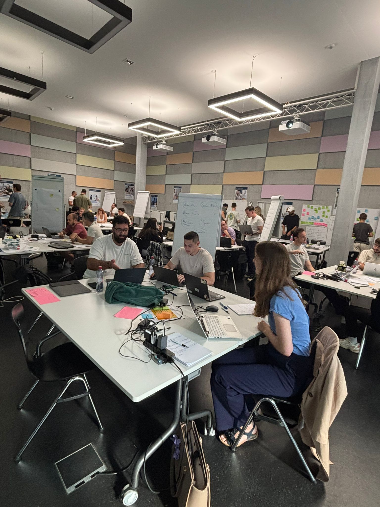 Grossraum mit mehreren Gruppentischen, im Vordergrund drei Teilnehmende an Laptops, dazwischen Post-its und ein beschriebenes Flipchart.