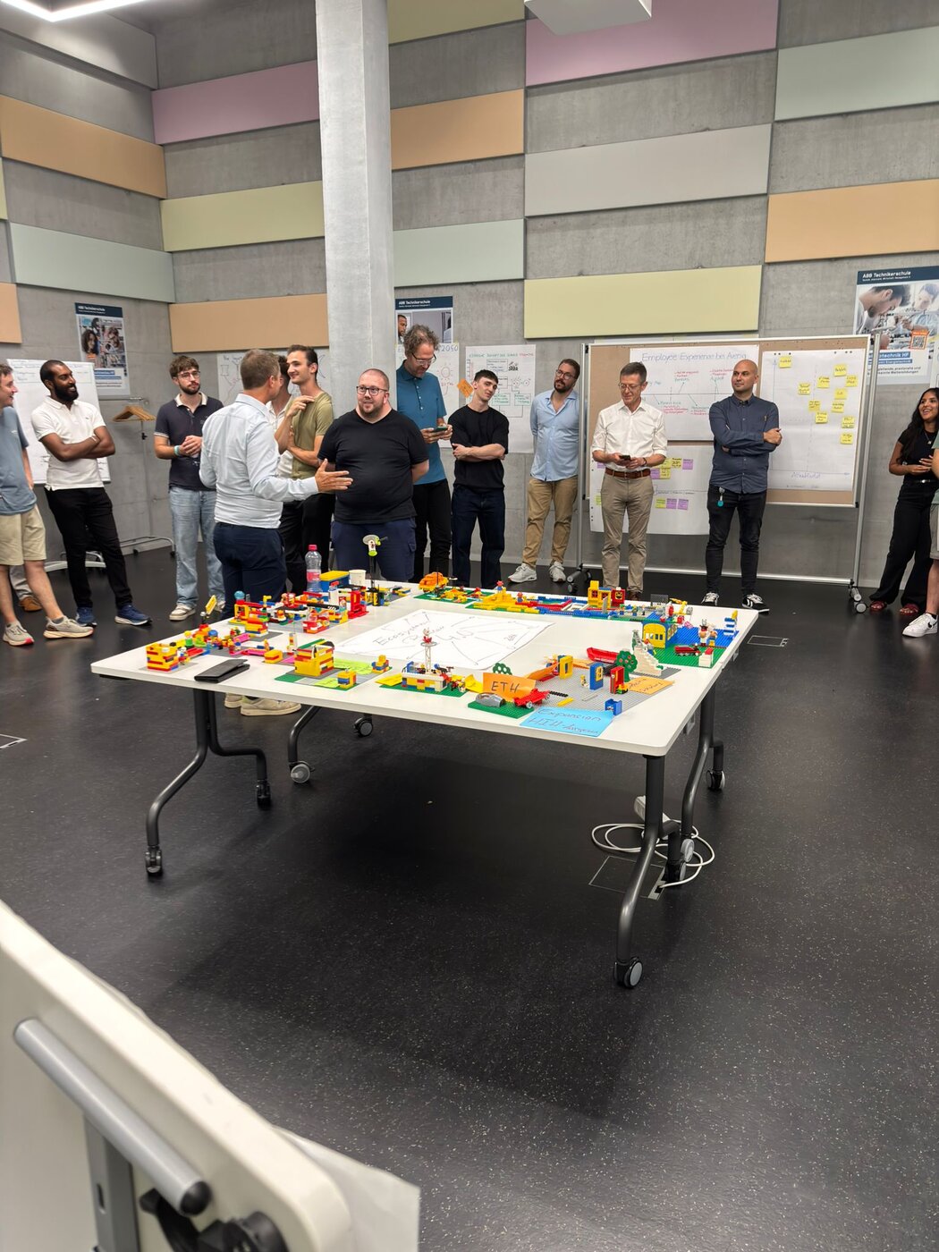 Teilnehmende und Firmenvertreter stehen im Kreis um einen Tisch, auf dem ein grosses Lego-Ökosystem aufgebaut ist, im Hintergrund Flipcharts und Poster.