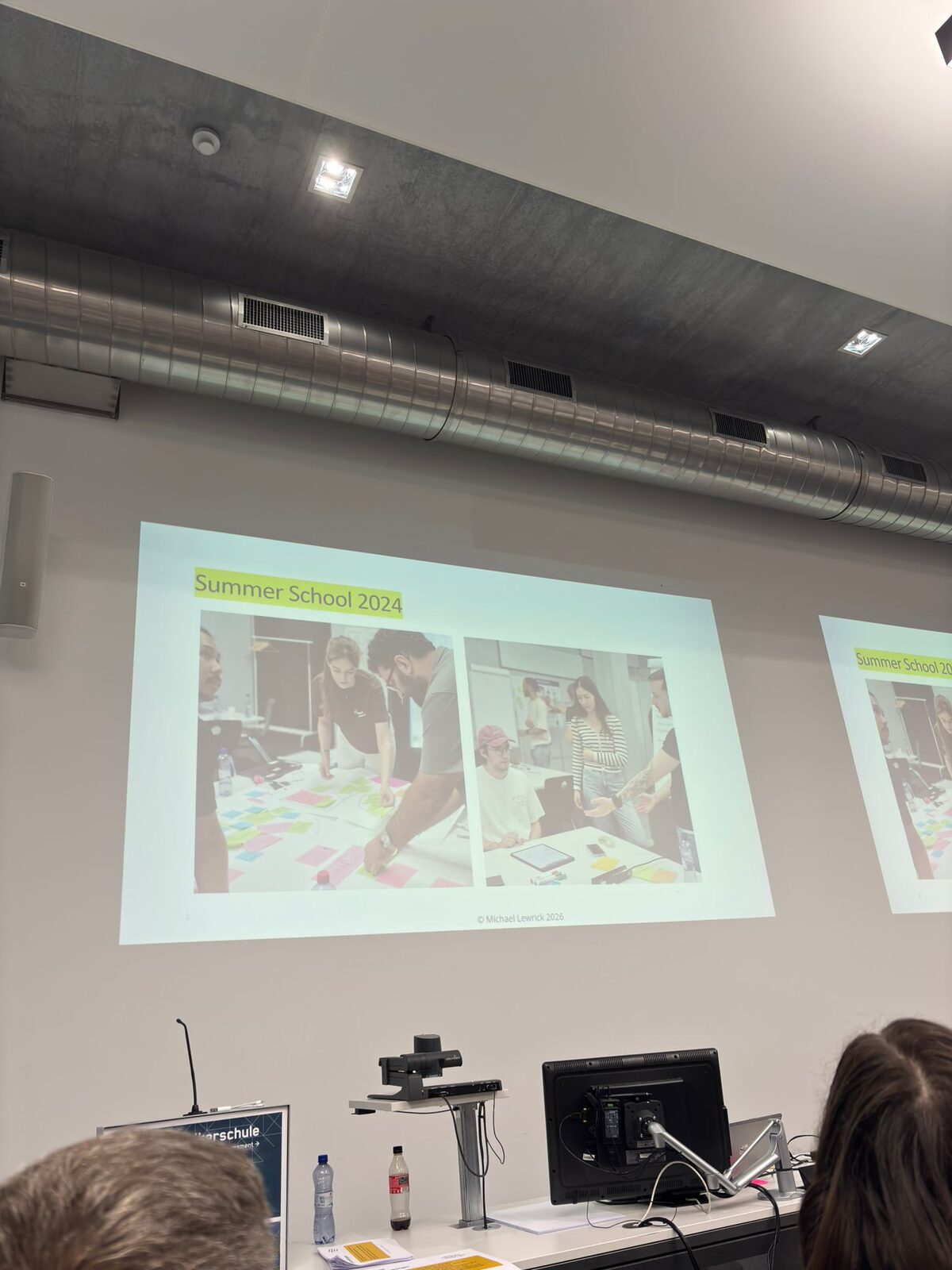 Projizierte Folie mit dem Titel Summer School 2024 und zwei Fotos von Teilnehmenden an einer Bodenmatrix aus Post-its.