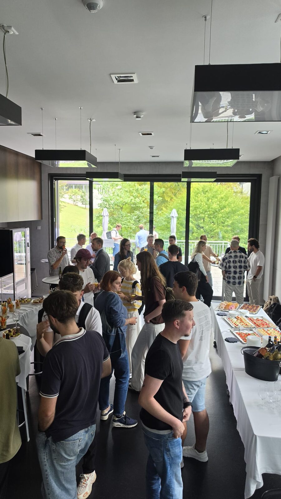 Teilnehmende stehen beim Apéro an Stehtischen und am Buffet, dahinter die Fensterfront ins Grüne.